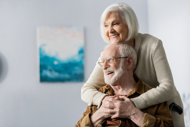 Χαρούμενη ηλικιωμένη γυναίκα που αγκαλιάζει ευτυχισμένο ανάπηρο σύζυγο  - Φωτογραφία, εικόνα