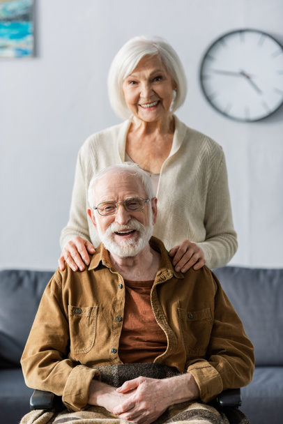 szczęśliwy starszy kobieta dotykając ramiona uśmiechnięty mąż siedzi na wózku inwalidzkim - Zdjęcie, obraz