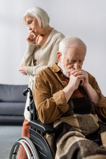 selective focus of sad senior wife standing behind disabled, ill man sitting in wheelchair with folded hands - Photo, Image