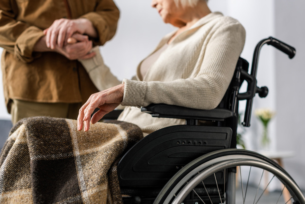 oříznutý pohled na staršího muže držícího za ruku postiženou, nemocnou ženu na invalidním vozíku - Fotografie, Obrázek