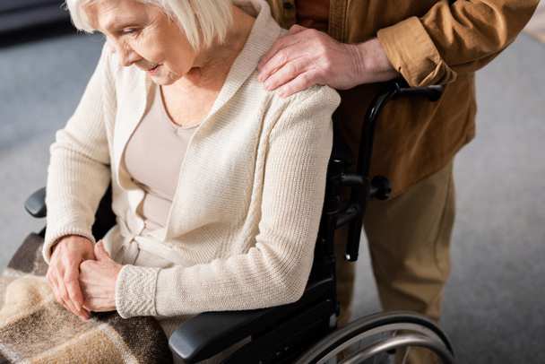 обрезанный вид пожилого мужчины, трогающего плечо жены-инвалида, сидящей в инвалидном кресле со склоненной головой
 - Фото, изображение