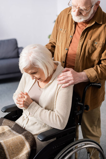 Yaşlı adam engelli karısının omzuna dokunuyor, bunamış, tekerlekli sandalyede başı eğik ve elleri sımsıkı oturuyor. - Fotoğraf, Görsel