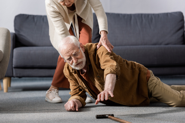 hombre mayor acostado en el suelo y tratando de conseguir bastón mientras su esposa le ayuda
 - Foto, Imagen
