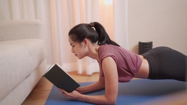 Ázsiai fit lány planking és olvasás könyv multitasking, önmotiváció zárlat alatt, otthoni edzés a nappaliban a kanapé mellett, otthoni nevelési iskola kilátás tanulmányi előadás, meleg fény - Felvétel, videó