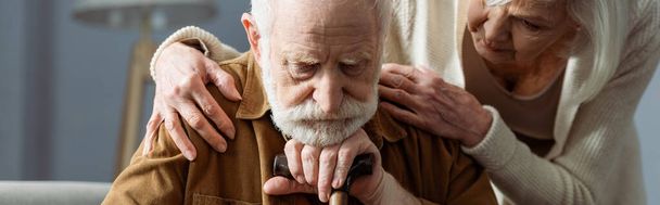 Panoramaaufnahme eines demenzkranken älteren Mannes, der mit Gehstock sitzt, während seine Frau ihn ansieht und seine Schultern berührt - Foto, Bild