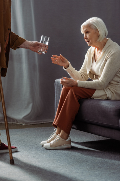 częściowy widok seniora dając szklankę wody do żony czuje się źle i siedzi na kanapie - Zdjęcie, obraz