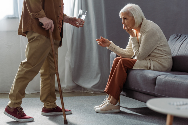 vista recortada de hombre mayor dando vaso de agua a la esposa sentirse mal y sentado en el sofá
 - Foto, Imagen