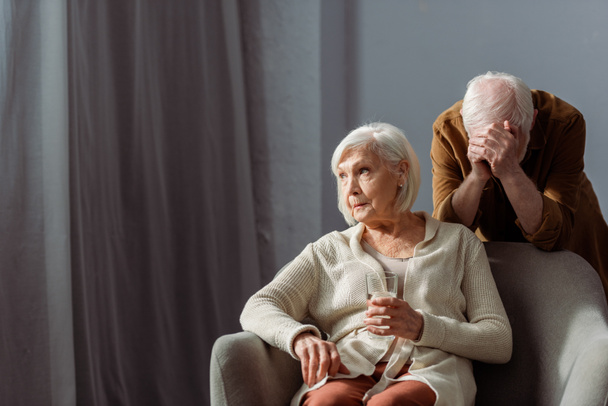 отчаявшийся пожилой человек, затемняющий лицо рукой, стоя позади больной жены, держа стакан воды
 - Фото, изображение