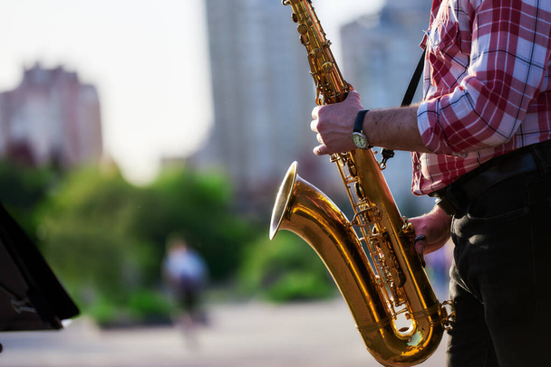 saxophonist plays a golden saxophone on the street with passers-by in sight. spring. musical reed wind instrument. tongue wooden brass instrument. bokeh. - Photo, Image