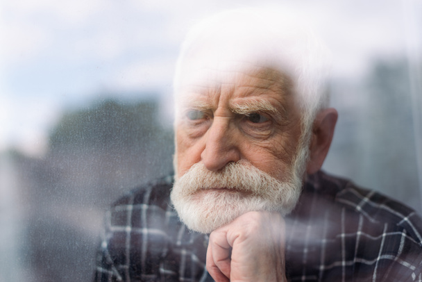 grieving senior man looking away through window glass while holding hand near chin - Fotó, kép