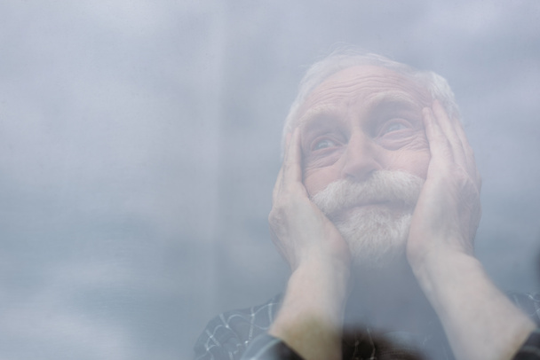 selective focus of senior, lonely man touching face and looking away through window glass - Photo, Image