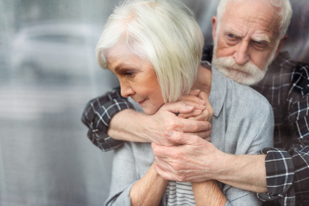 засмучений старший чоловік обіймає дружину, хворий на деменцію, стоячи біля вікна
 - Фото, зображення