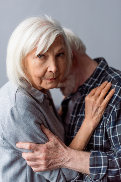 αναστατωμένη ηλικιωμένη γυναίκα που κοιτάζει την κάμερα ενώ αγκαλιάζει τον σύζυγο που πάσχει από άνοια - Φωτογραφία, εικόνα