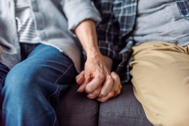 обрезанный вид пожилой пары, сидящей на диване и держащейся за руки
  - Фото, изображение