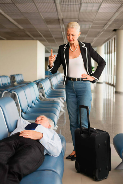 Businessman asleep on seat in airport lounge, businesswoman standing over him pointing finger - Фото, изображение