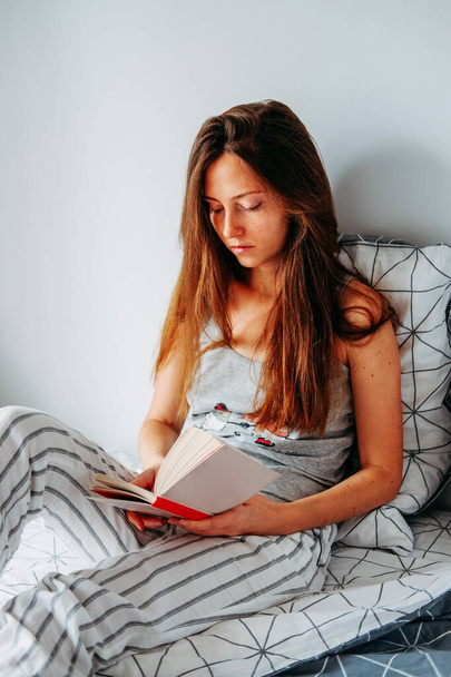 若い赤毛の女性がベッドで読んでいる。遅い朝のルーチン. - 写真・画像