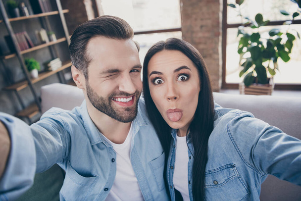 Close up photo of carefree playful positive two people man woman travel trip make selfie show tongue out wink blink sit couch in house indoors - Foto, Imagem