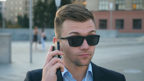 Camera spinning around of handsome businessman talking on phone outdoor. Young business man in sunglasses having conversation on cellphone. Portrait of guy speaking on smartphone outside. Slow motion - Video