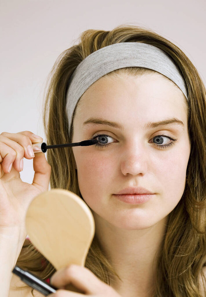Girl applying mascara while looking at hand mirror - Fotografie, Obrázek