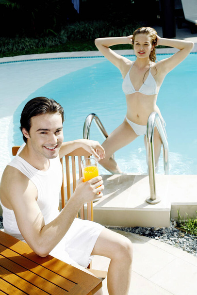 Man holding a glass of orange juice with his girlfriend climbing up from the swimming pool - Фото, зображення