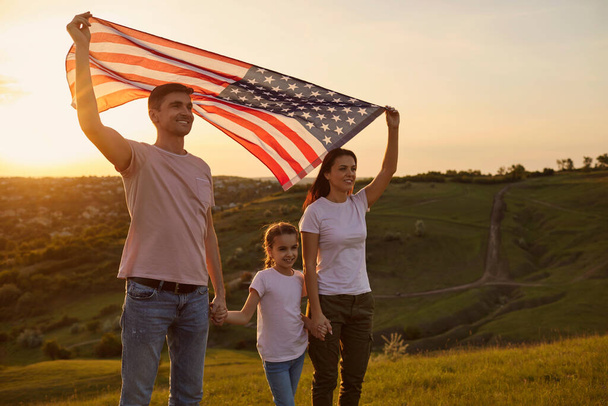 Young parents with their daughter holding American flag in countryside at sunset. Independence Day celebration - Zdjęcie, obraz