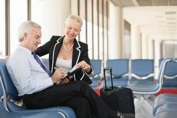 Businessman writing in organizer, businesswoman sitting beside him in airport lounge - Valokuva, kuva