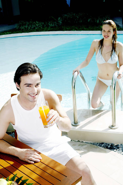 Man holding a glass of orange juice with his girlfriend climbing up from the swimming pool - Foto, Imagem