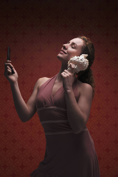 Woman holding seashell to ear while holding cell phone - Foto, afbeelding