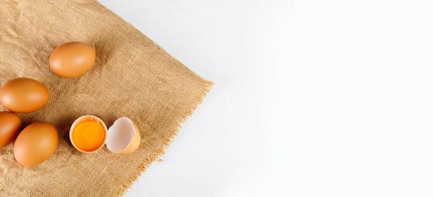 Top view broken raw egg with the yolk and albumen on a linen tablecloth on white background. Raw eggs on white table with copy space banner. - Fotoğraf, Görsel