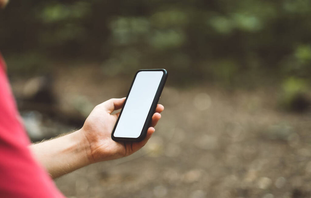 A man holds a smartphone with a white screen in a forest clearing while hiking. High quality photo - Photo, image