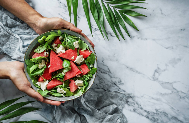Mains tenant une salade de pastèque fraîche d'été saine avec roquette, épinards et légumes verts sur fond de marbre clair avec des feuilles tropicales. Nourriture saine, manger propre, salade bol Bouddha, vue sur le dessus - Photo, image