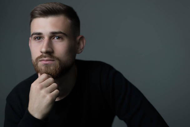 pensive young man of twenty five years old. A bearded man thinking, holding his hand to his chin, in black. Studio photo on a gray background. A philosopher, thinking, a man thinks, a look, a portrait of a man. - Photo, Image