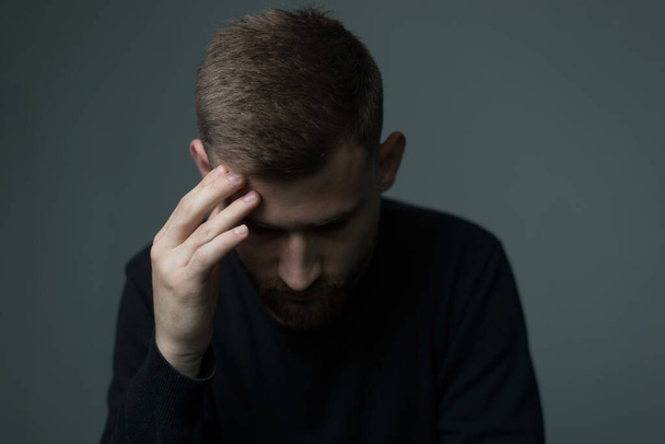 Portrait of a bearded young man of twenty-five, in black, holds his hand to his head, suffers from a headache. On a gray background. Depressed state, unhealthy guy, headache, migraine, mental health, impaired condition, weakness, dizziness - Photo, Image