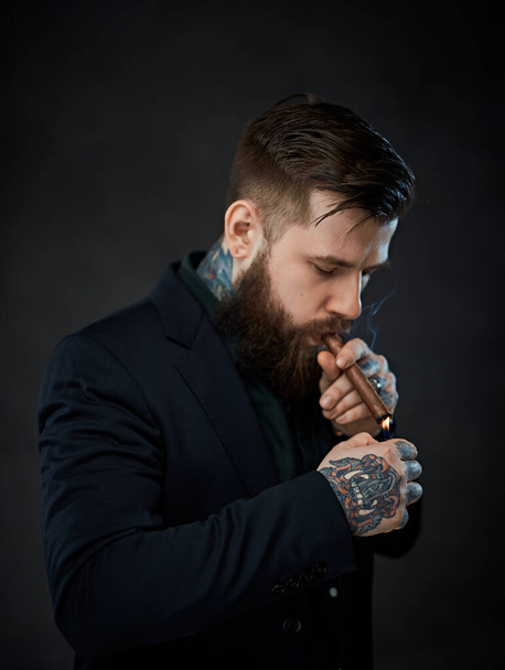 Elegantly dressed bearded male with tattoos on his neck and hand lights a cigar - Fotó, kép