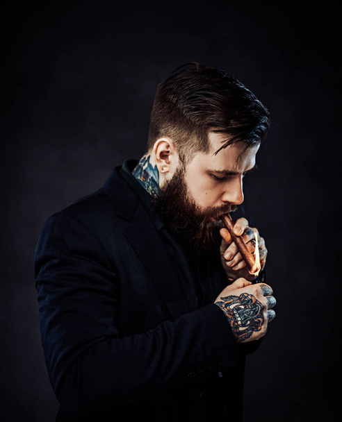 Elegantemente vestido barbudo macho com tatuagens em seu pescoço e mão acende um charuto
 - Foto, Imagem