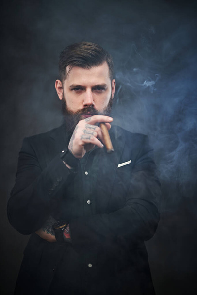 Brutal young businessman holds a cigar and looks at the camera in a smoky dark studio - Fotoğraf, Görsel