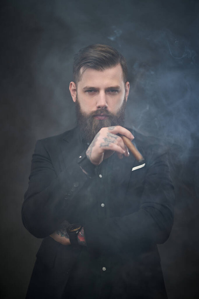 Brutal jovem empresário segura um charuto e olha para a câmera em um estúdio escuro fumegante
 - Foto, Imagem