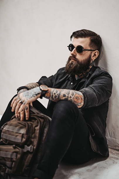 Bearded hipster guy in sunglasses with backpack sitting on a floor in studio - Foto, Imagem