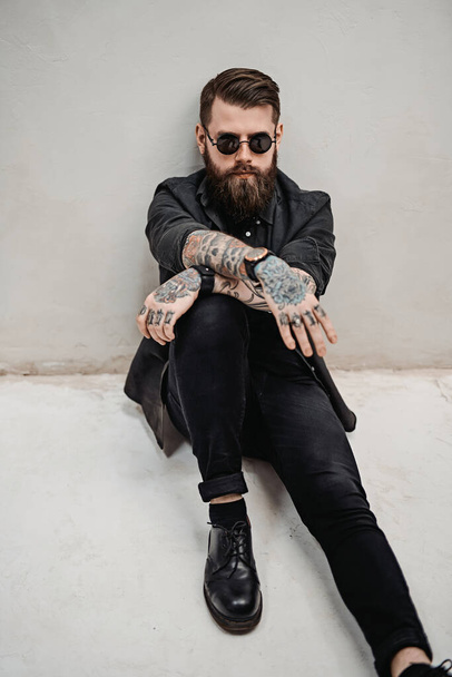 Cool bearded hipster guy in sunglasses sitting of a floor in studio - Foto, afbeelding