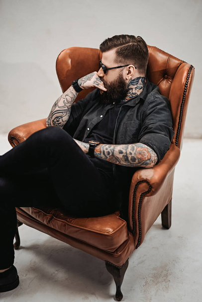 Cool barbudo jovem sentado em uma cadeira vintage em um estúdio
 - Foto, Imagem