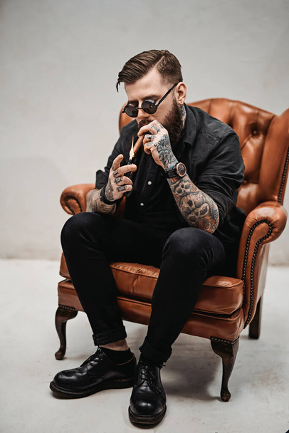 Прохолодний татуйований бородатий чоловік запалює сигару, сидячи на старовинному стільці
 - Фото, зображення