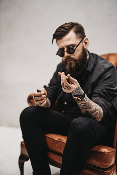 Cool tattooed bearded man lights a cigar while sitting on a vintage chair - Φωτογραφία, εικόνα