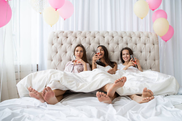 Pajama party. Attractive young smiling women in pajamas drinking champagne while having a slumber party in the bedroom. - Photo, Image