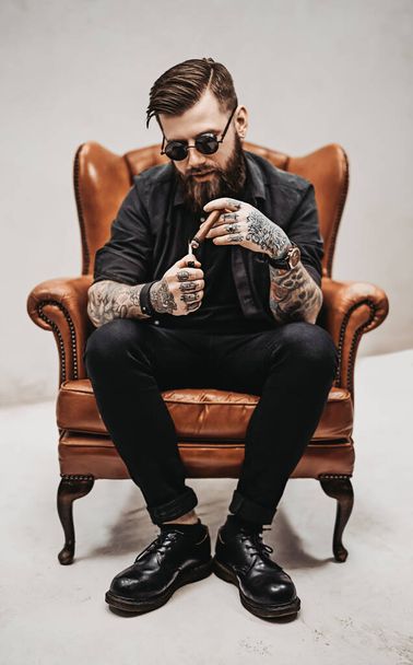 Hombre barbudo tatuado con corte de pelo elegante en gafas de sol que fuma un cigarro mientras está sentado en una silla vintage en el estudio
 - Foto, Imagen