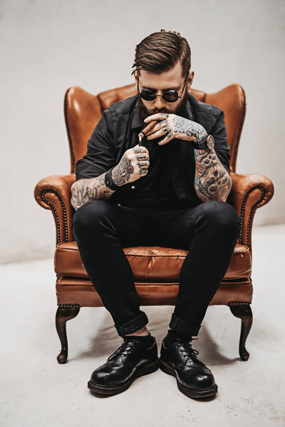 Tattooed bearded man with stylish haircut in sunglasses who smokes a cigar while sitting on a vintage chair in studio - 写真・画像