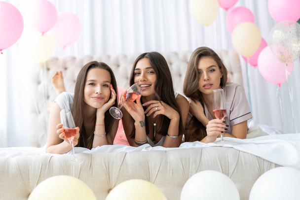 Πάρτι πυτζάμα. Ελκυστικές νεαρές χαμογελαστές γυναίκες με πιτζάμες πίνοντας σαμπάνια ενώ κάνουν πιτζάμα πάρτι στην κρεβατοκάμαρα. - Φωτογραφία, εικόνα