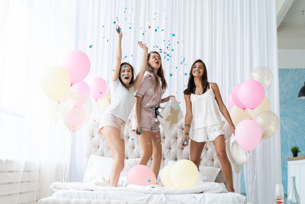 Four attractive young women in pajamas smiling and gesturing while jumping in bedroom with confetti flying everywhere - Photo, image