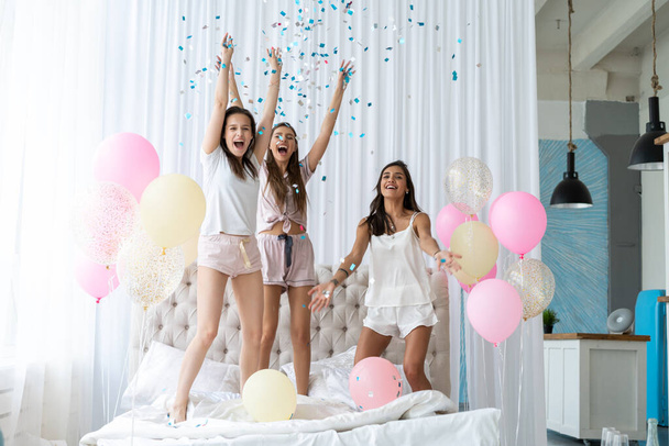 Four attractive young women in pajamas smiling and gesturing while jumping in bedroom with confetti flying everywhere - Fotó, kép