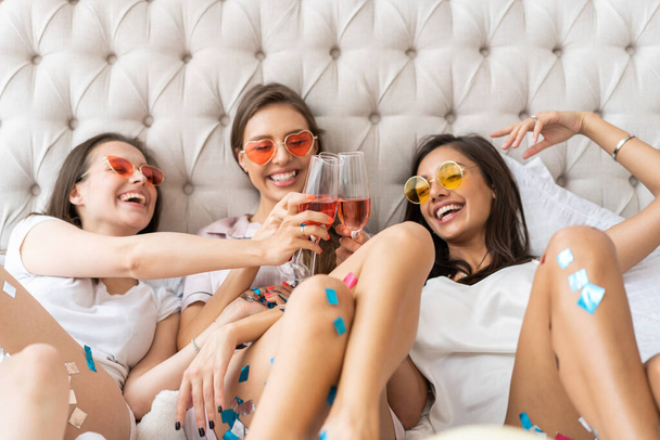 Pajama party. Attractive young smiling women in pajamas drinking champagne while having a slumber party in the bedroom. - Photo, image