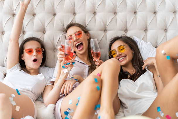 Pajama party. Attractive young smiling women in pajamas drinking champagne while having a slumber party in the bedroom. - Photo, image